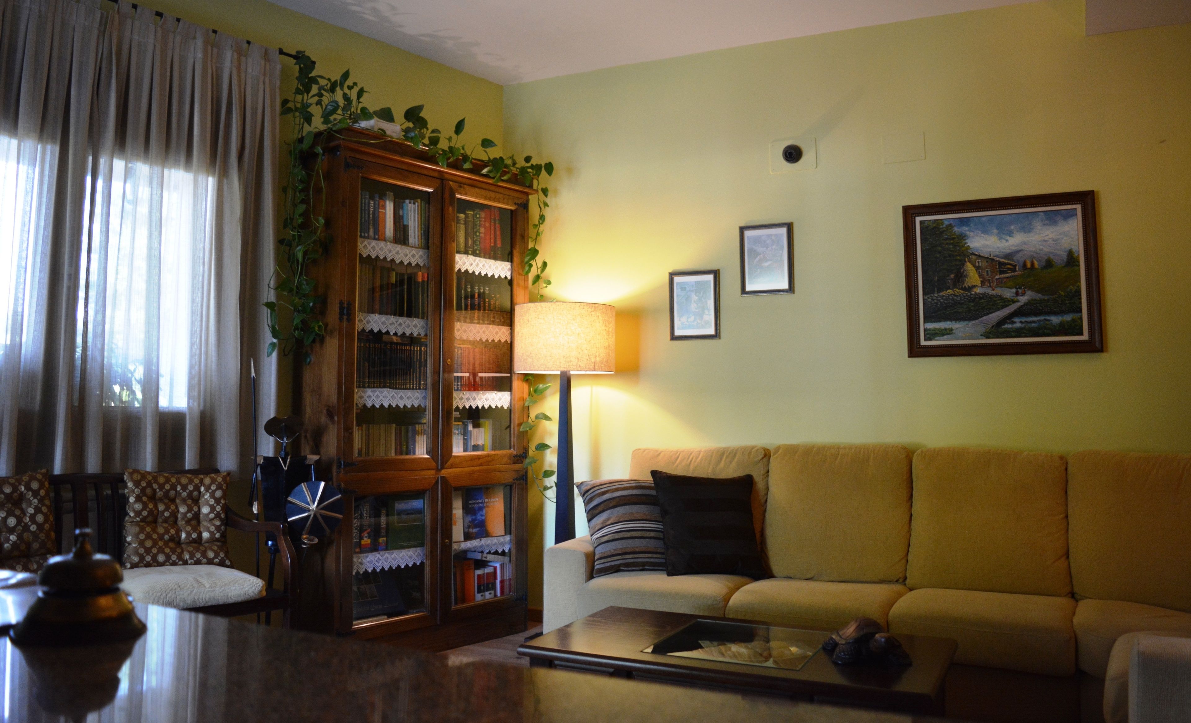 Una sala de estar con un sofá amarillo, una estantería de madera, una lámpara y un cuadro en la pared.