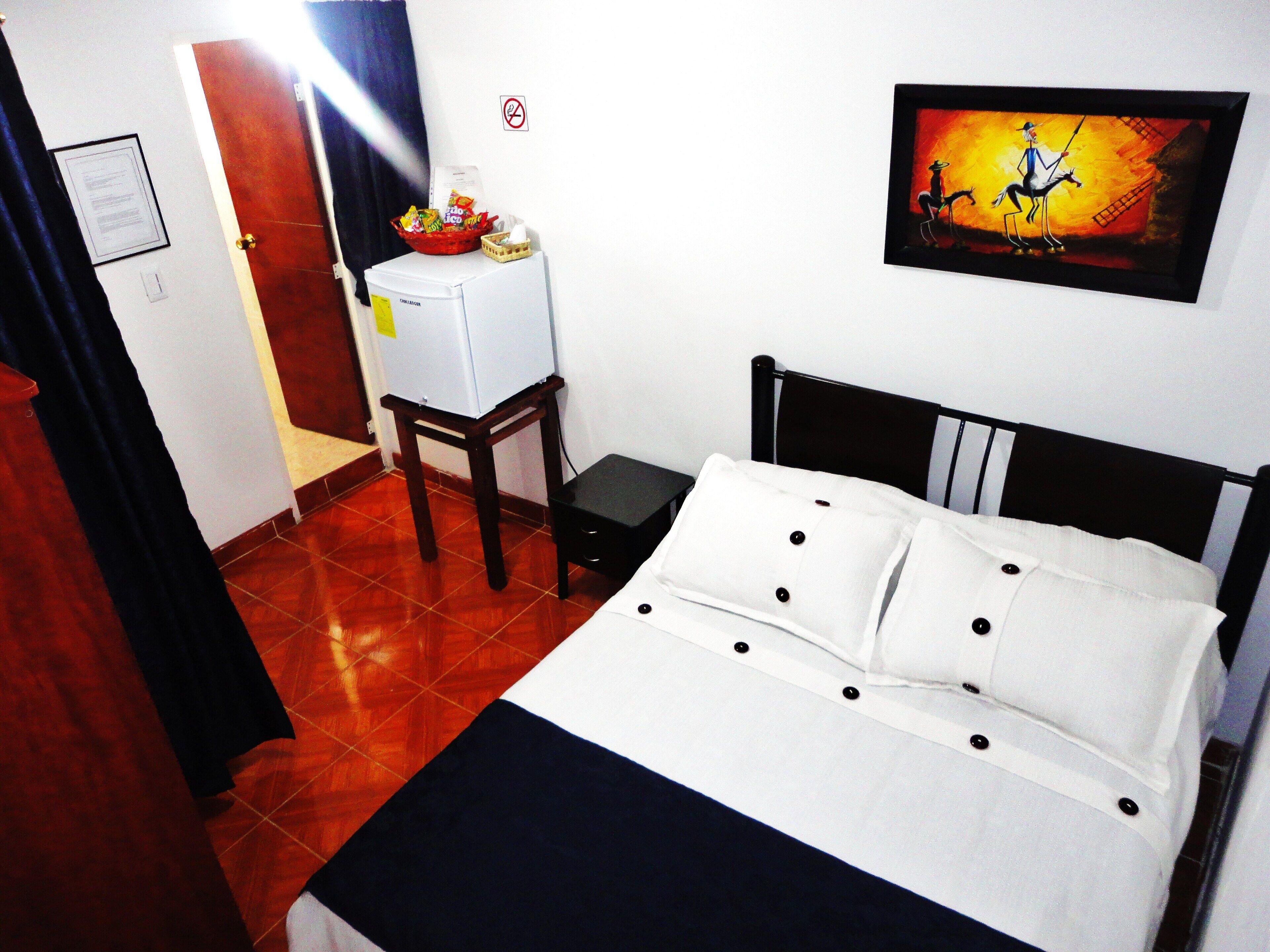 A hotel room with a bed, a small table, a refrigerator, and a painting on the wall.