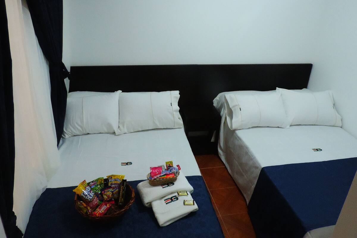 Une chambre d’hôtel avec deux lits simples, chacun étant équipé de draps et d’oreillers blancs. Un panier de collations se trouve sur le côté gauche du lit, et un ensemble de serviettes sur le côté droit.