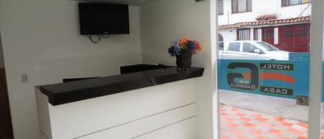 A hotel reception area with a flat-screen TV mounted on the wall, a reception desk, and a vase with flowers.