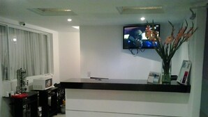 Ein moderner Empfangsbereich mit Wand-Fernseher, einem schwarzen Empfangstisch und einer Vase mit Blumen auf der rechten Seite.