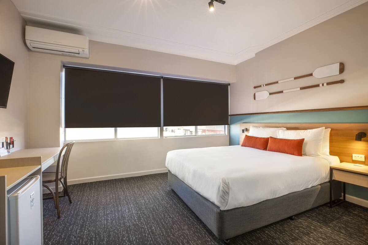 A hotel room with a bed, desk, chair, and a large window with blinds.