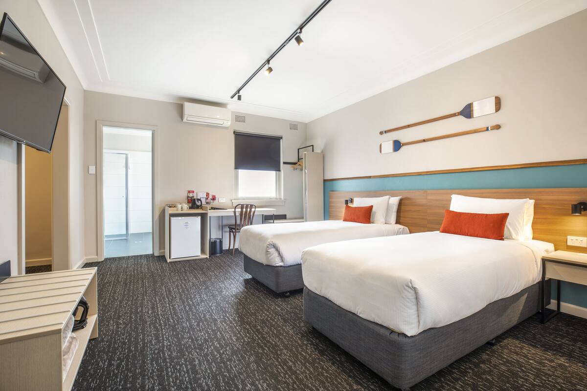 A hotel room with two beds, a desk, a chair, a TV, and a wall-mounted shelf with items.