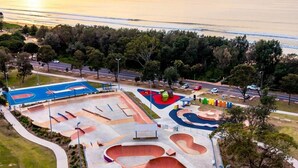 Vista aérea de un parque de skate con diversas rampas y una cancha de baloncesto, rodeado de árboles y una carretera.