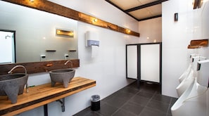 Een moderne badkamer met twee granieten wasbakken, een houten wastafelmeubel, een spiegel, een zeepdispenser en een urinoir.