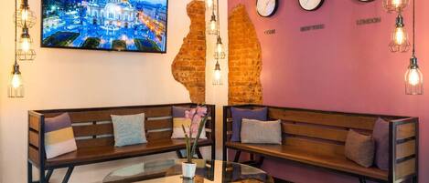 A lobby with two wooden benches, a glass coffee table, a large cityscape painting, a brick wall, and clocks on the wall.