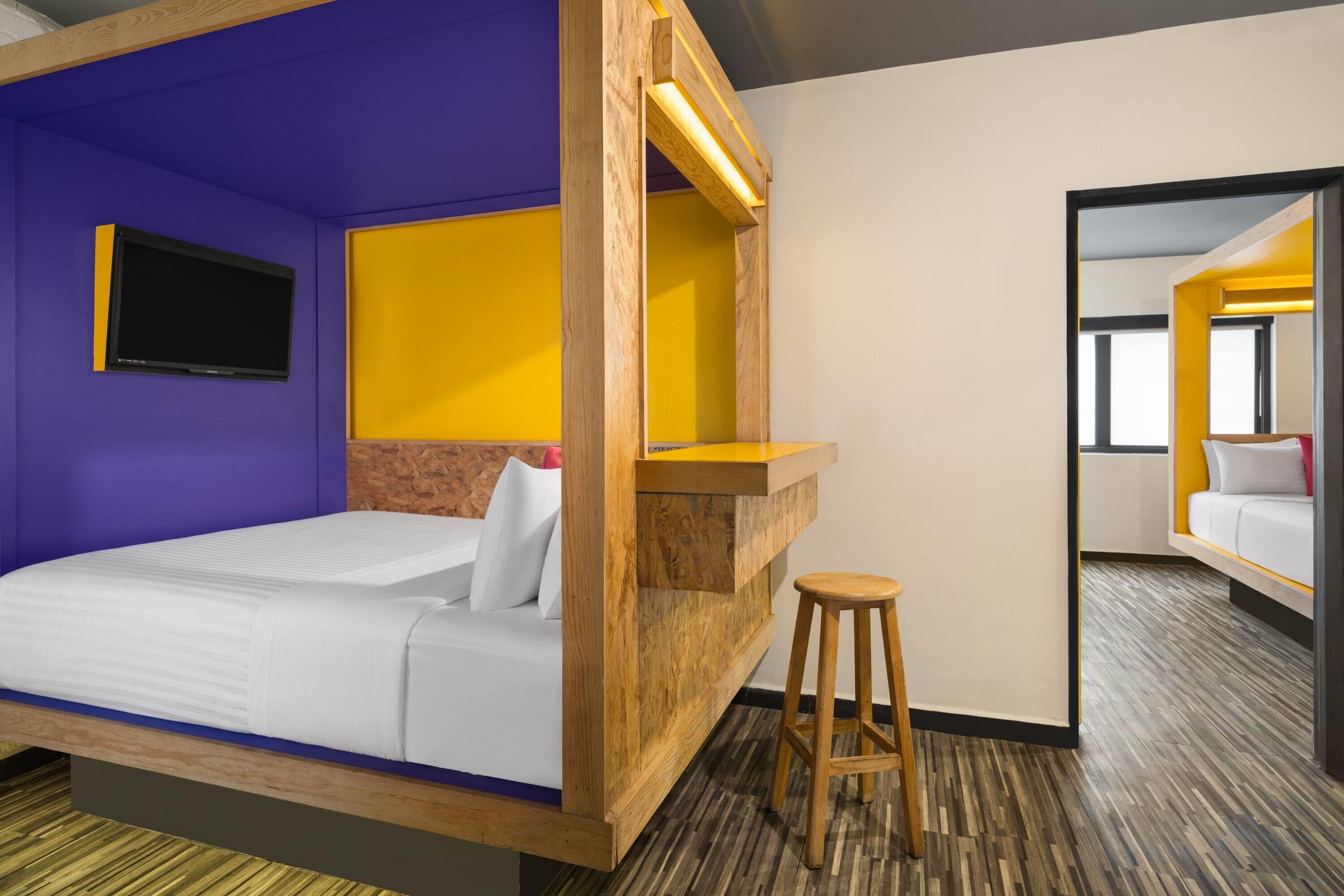 A hotel room with a bed, a small stool, a wooden headboard, a mirror, and a television.