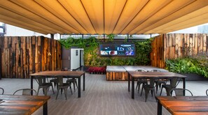 An outdoor patio with wooden tables and chairs, a large TV screen, and a wooden fence with greenery. - Hotel MX mas roma CDMX, Trademark Collection by Wyndham (Mexico City)