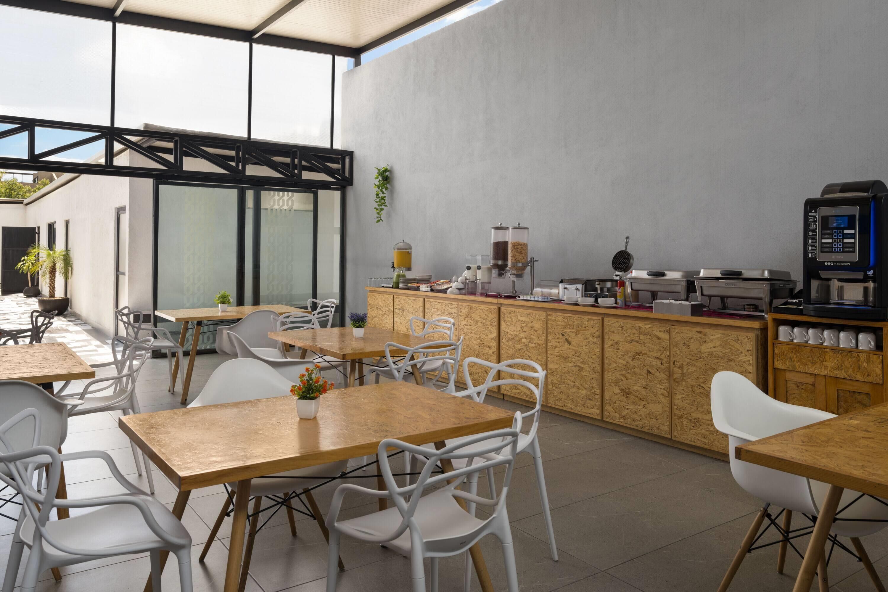 Un café moderno con mesas de madera y sillas blancas, una máquina de café y un dispensador de zumo.