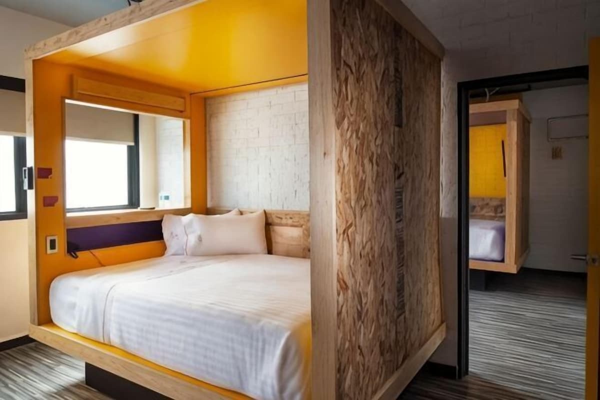 Quarto de hotel moderno com cama, cabeceira de madeira, um espelho grande e vista para o exterior.