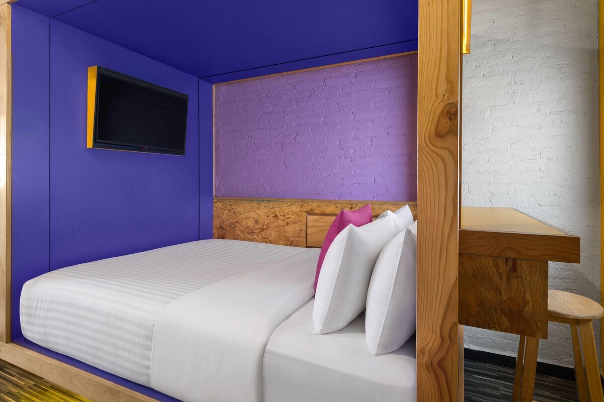 Une chambre avec un lit, une tête de lit en bois, une petite table et un téléviseur fixé au mur.