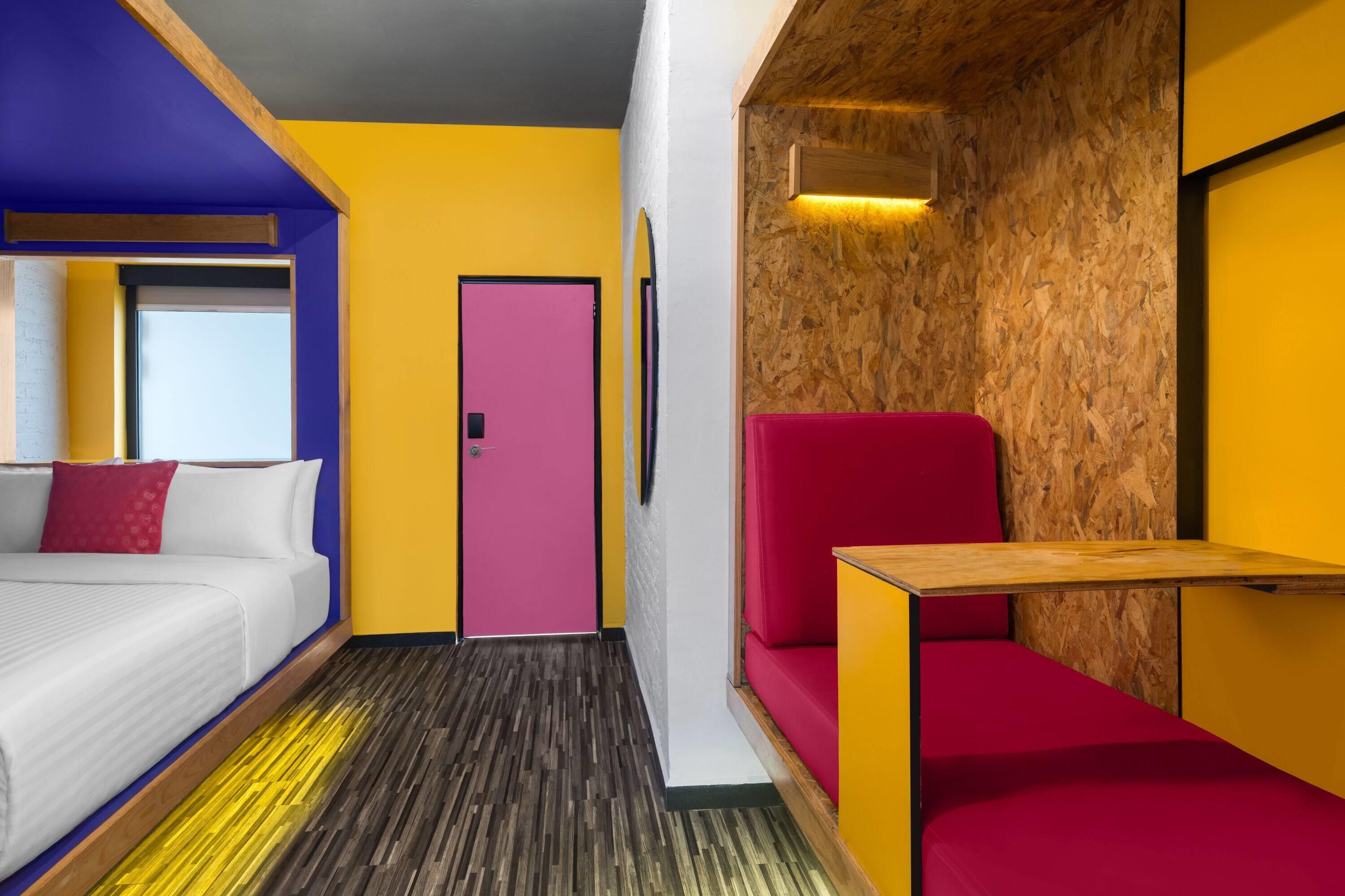 A hotel room with a bed, a red chair, a wooden table, and a door.