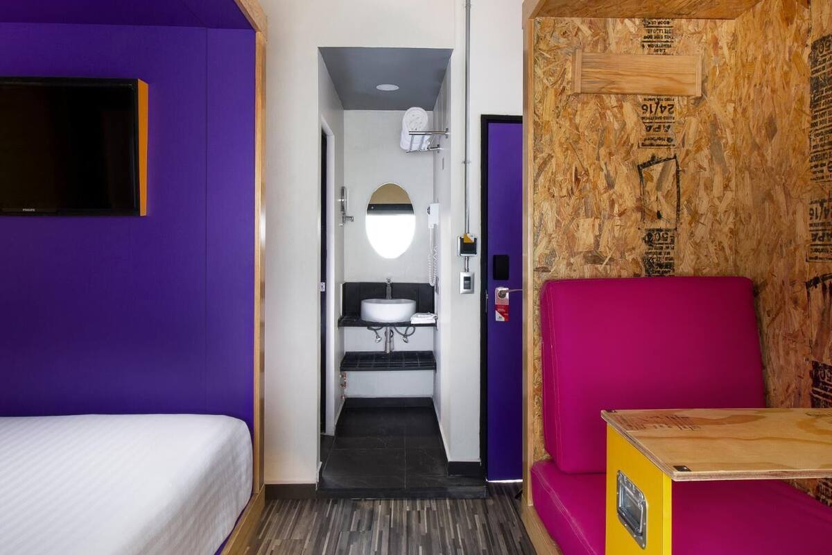Kompakti hotellihuone, jossa on sänky, pieni työpöytä, kylpyhuone, jossa on pesuallas ja peili, sekä violetti seinä.