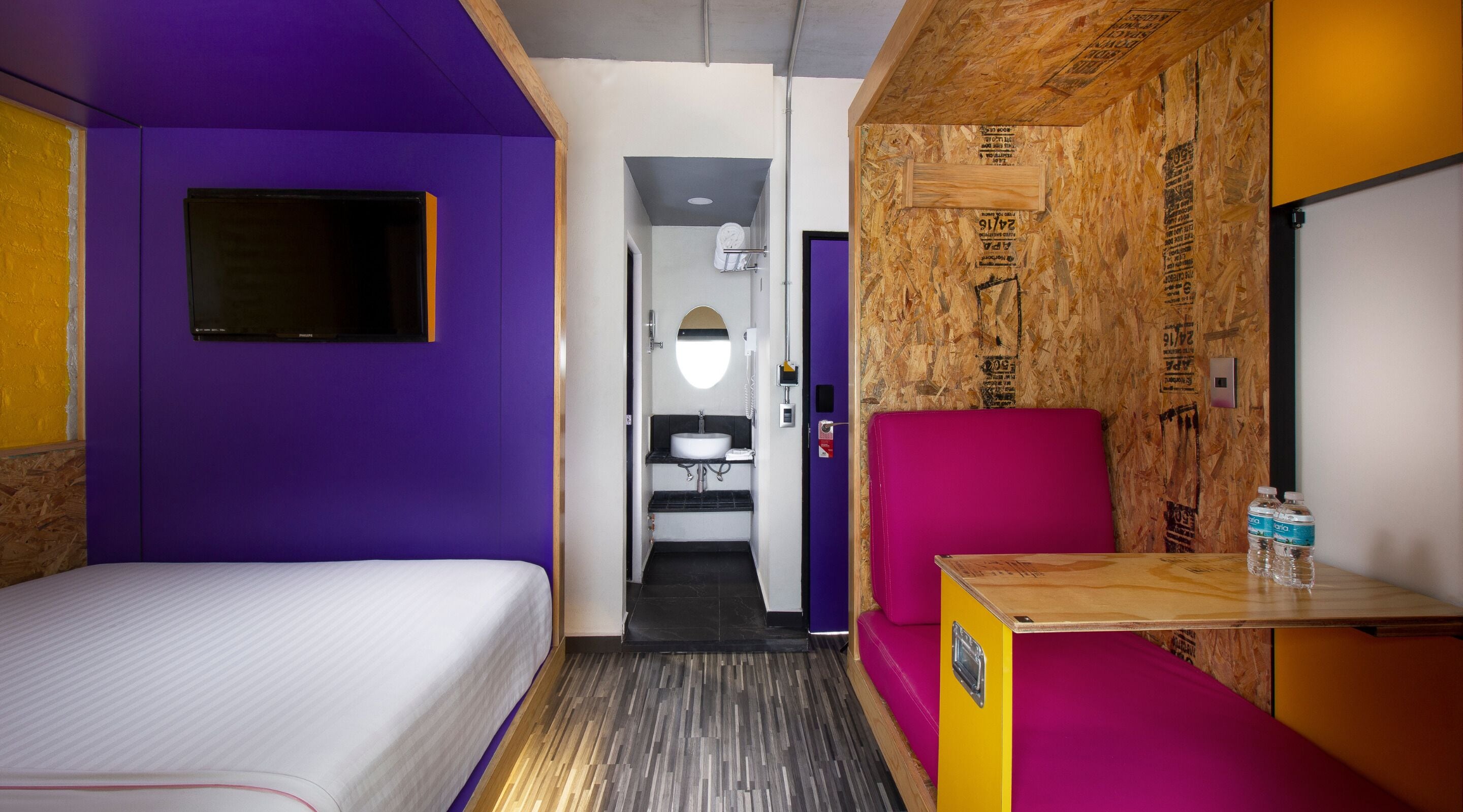Habitación de hotel compacta con una cama, una mesita con botellas de agua, un sillón morado y un baño con lavamanos y espejo.