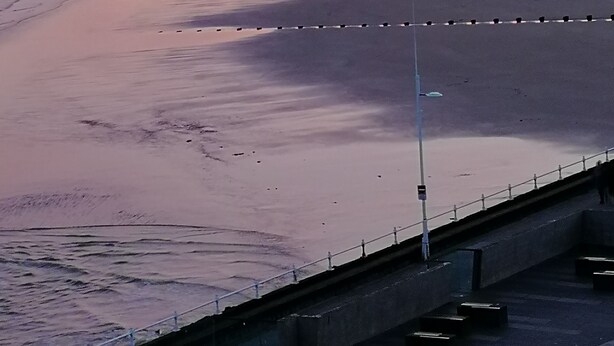 A beach with a calm sea, a few people walking, and wind turbines in the distance. - Rowntree Holiday Flats (Bridlington)