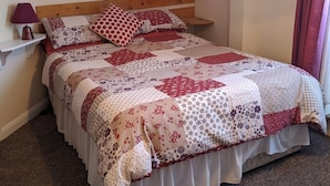 A neatly made bed with a floral-patterned quilt, a bedside table with a lamp, and a wall-mounted picture frame. - Rowntree Holiday Flats (Bridlington)
