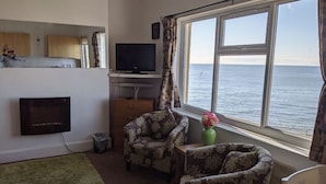 A living room with a television, two patterned armchairs, a small table, and a view of the sea through a large window. - Rowntree Holiday Flats (Bridlington)