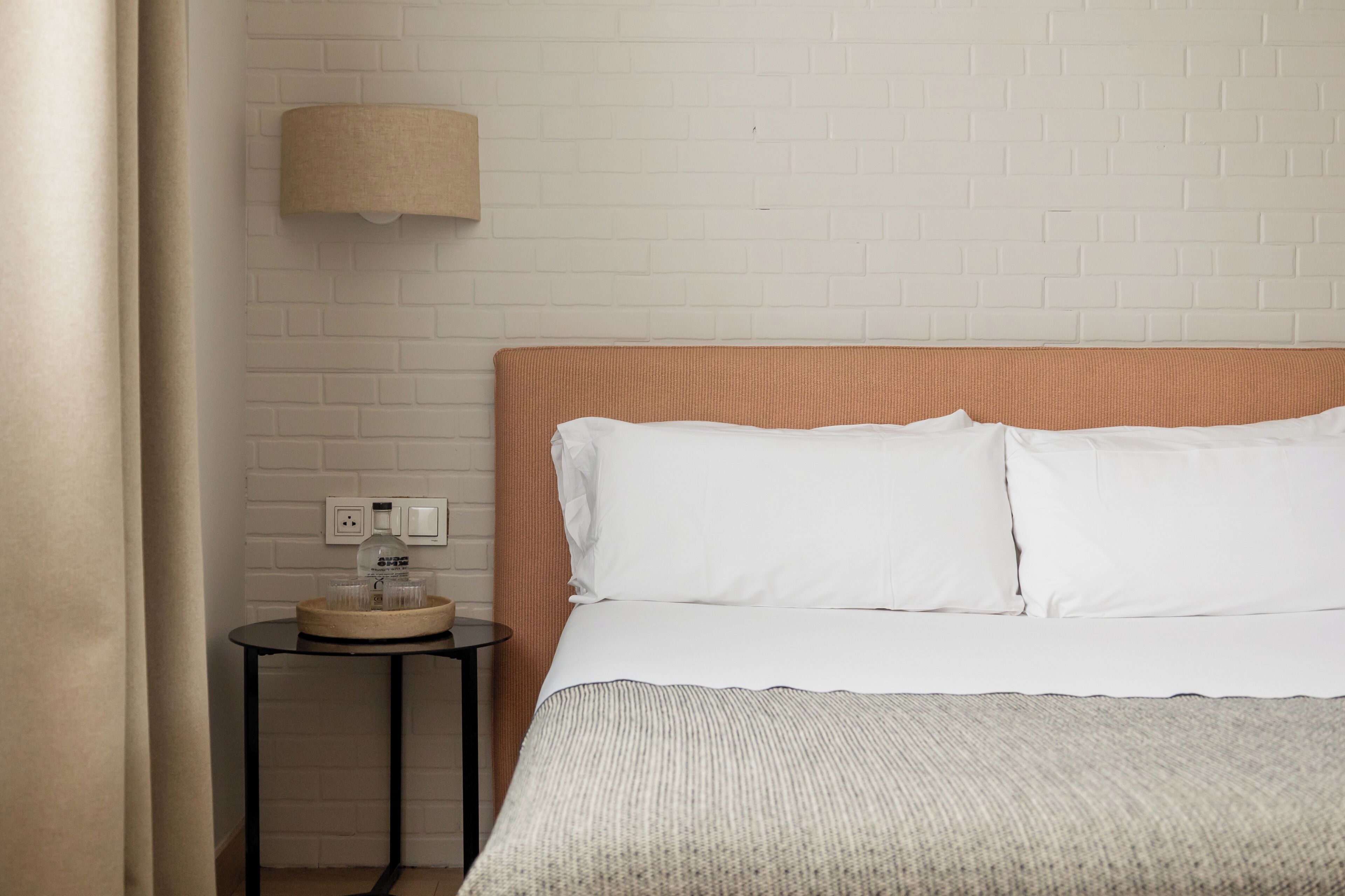 A hotel room with a white brick wall, a wooden headboard, a round table with a lamp, and a bed with white bedding.