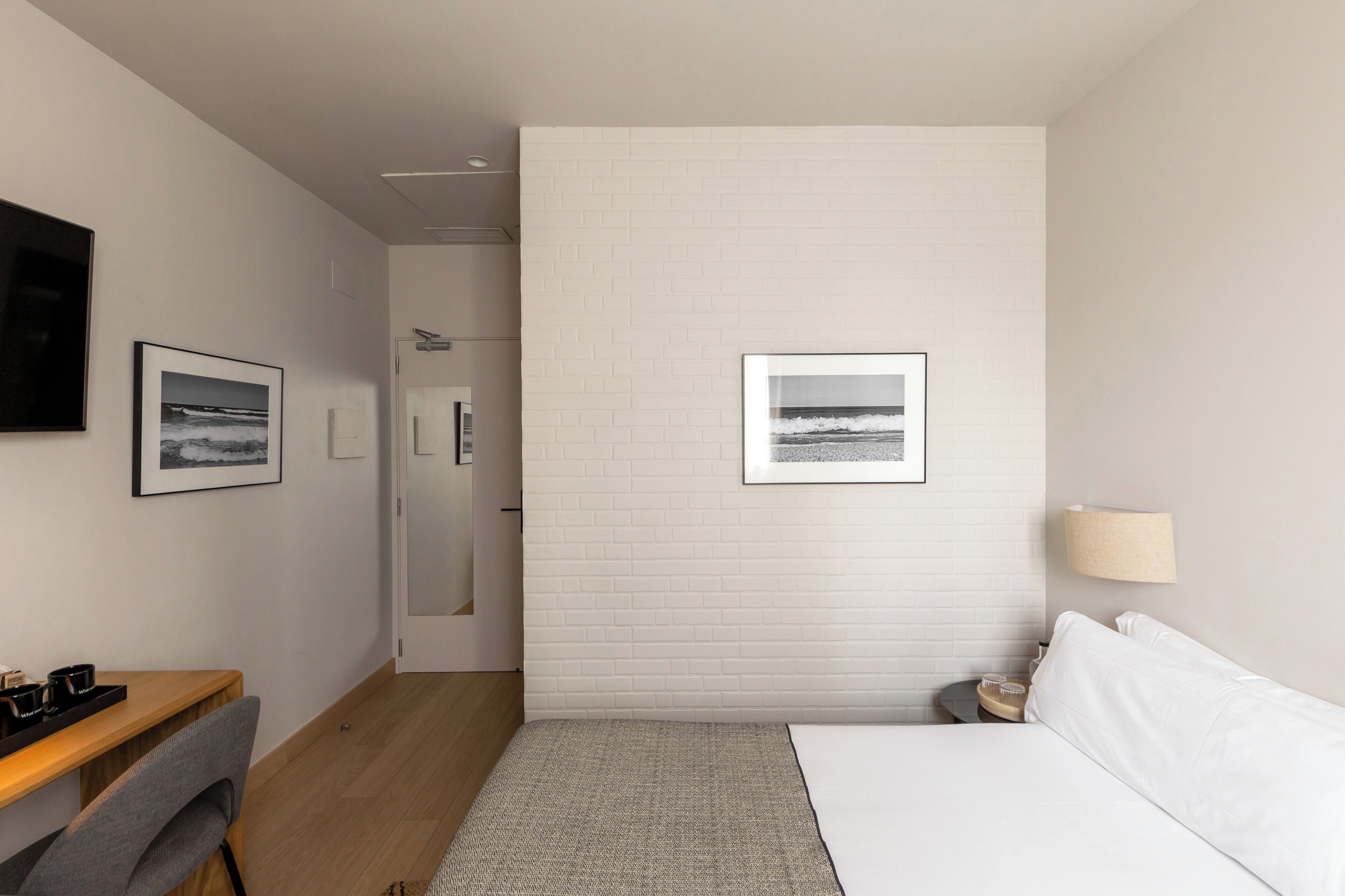 A hotel room with a bed, a desk, a chair, a lamp, and two framed pictures on the wall.