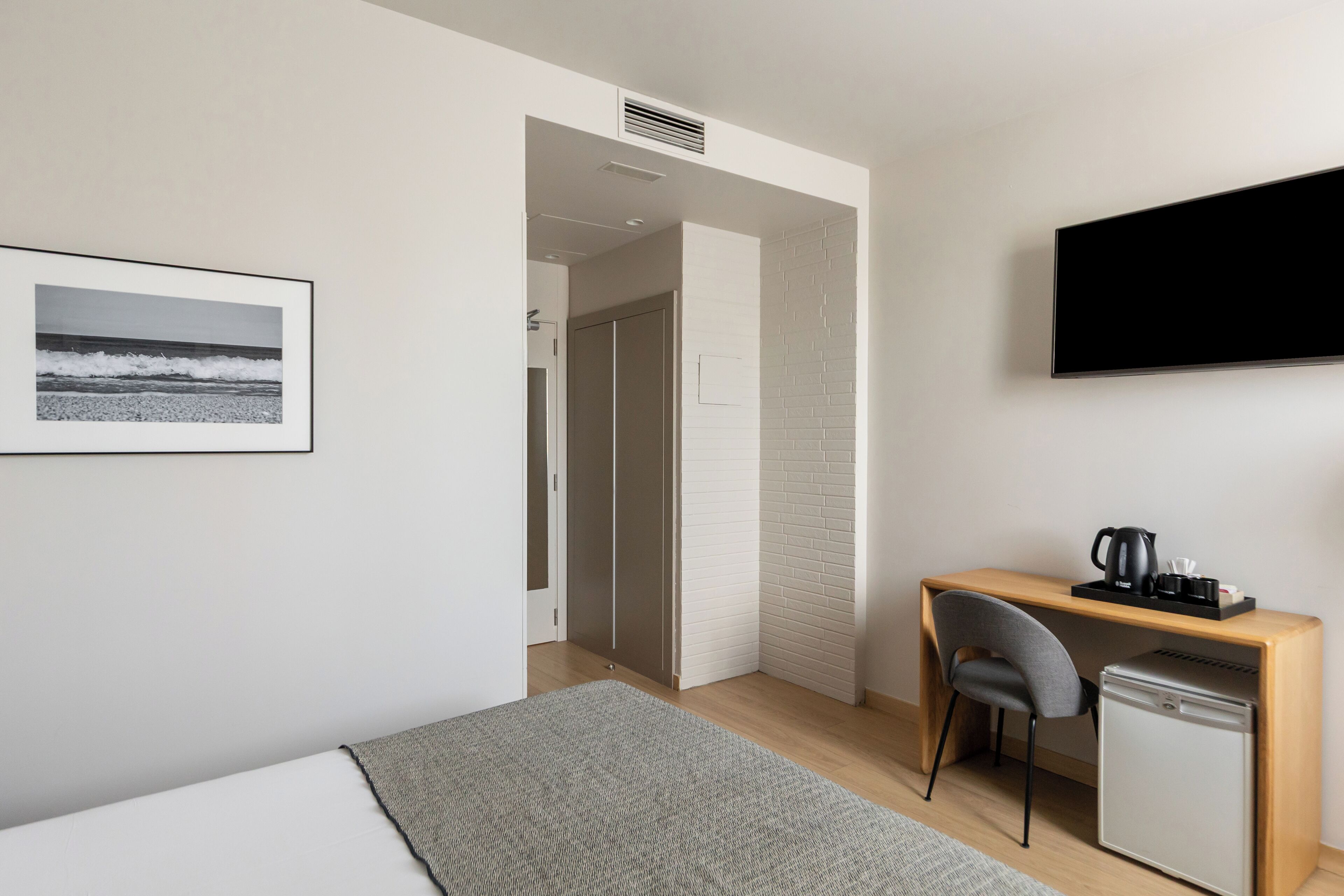 Una habitación de hotel moderna con una cama, un escritorio con una silla, un refrigerador pequeño, un televisor y una imagen del océano en la pared.