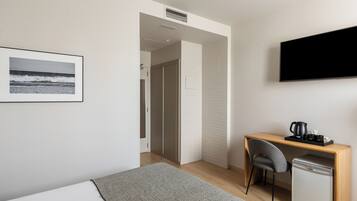 Una habitación de hotel moderna con una cama, un escritorio con una silla, un refrigerador pequeño, un televisor y una imagen del océano en la pared.