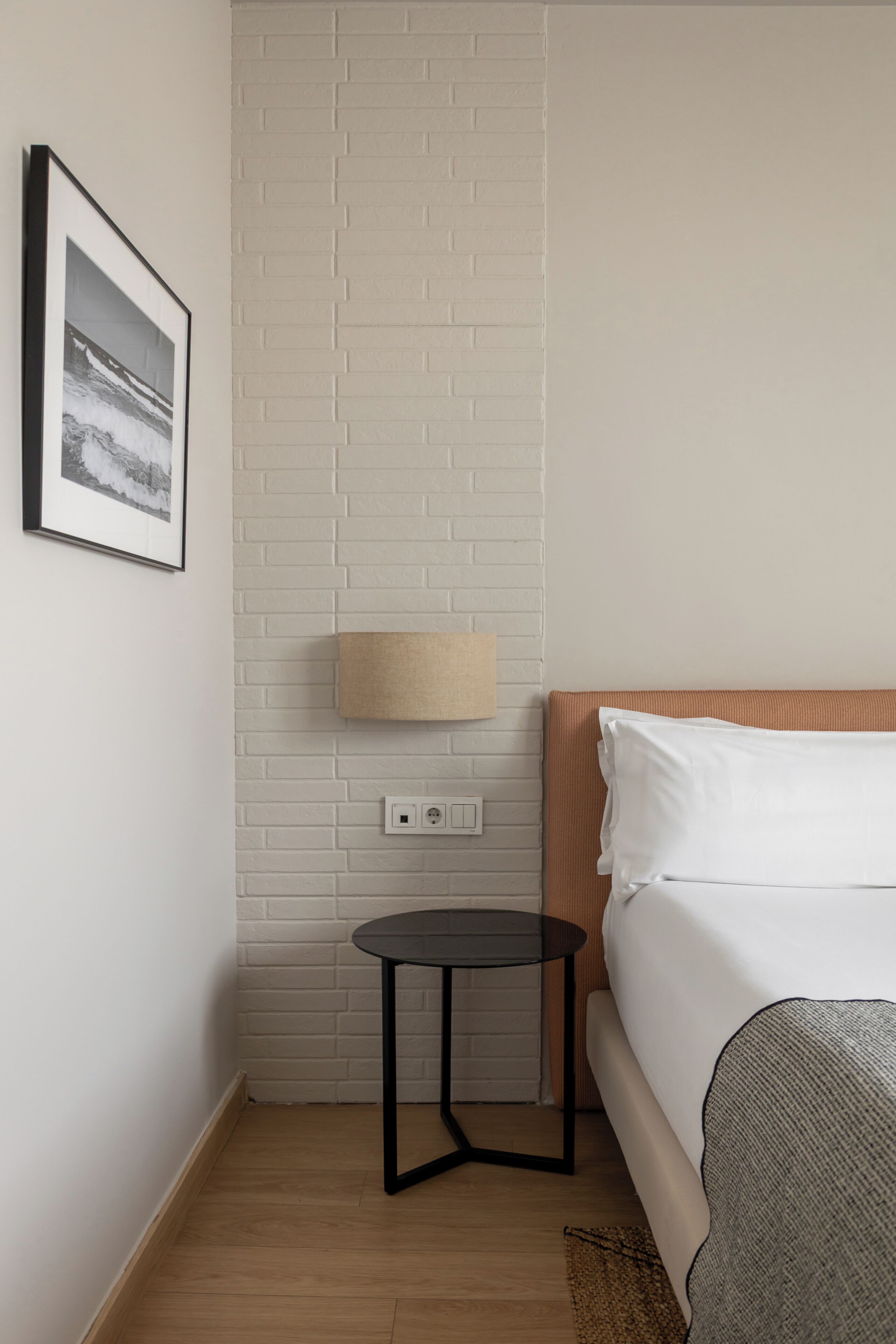 Una habitación de hotel con pared de ladrillo blanco, una mesa redonda negra, una cama con ropa de cama blanca y un cuadro en la pared.