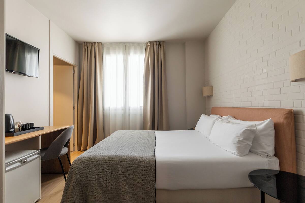 A hotel room with a bed, a desk with a chair, a television, and a window with curtains.