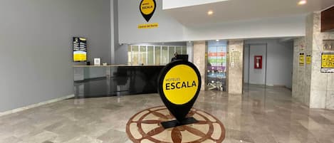 Un vestíbulo de hotel con un mostrador de recepción, un gran letrero amarillo que indica 'HOTELES ESCALA Central del Norte' y un diseño de piso circular.