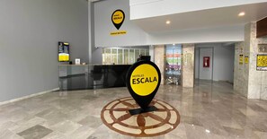 Un vestíbulo de hotel con un mostrador de recepción, un gran letrero amarillo que indica 'HOTELES ESCALA Central del Norte' y un diseño de piso circular.