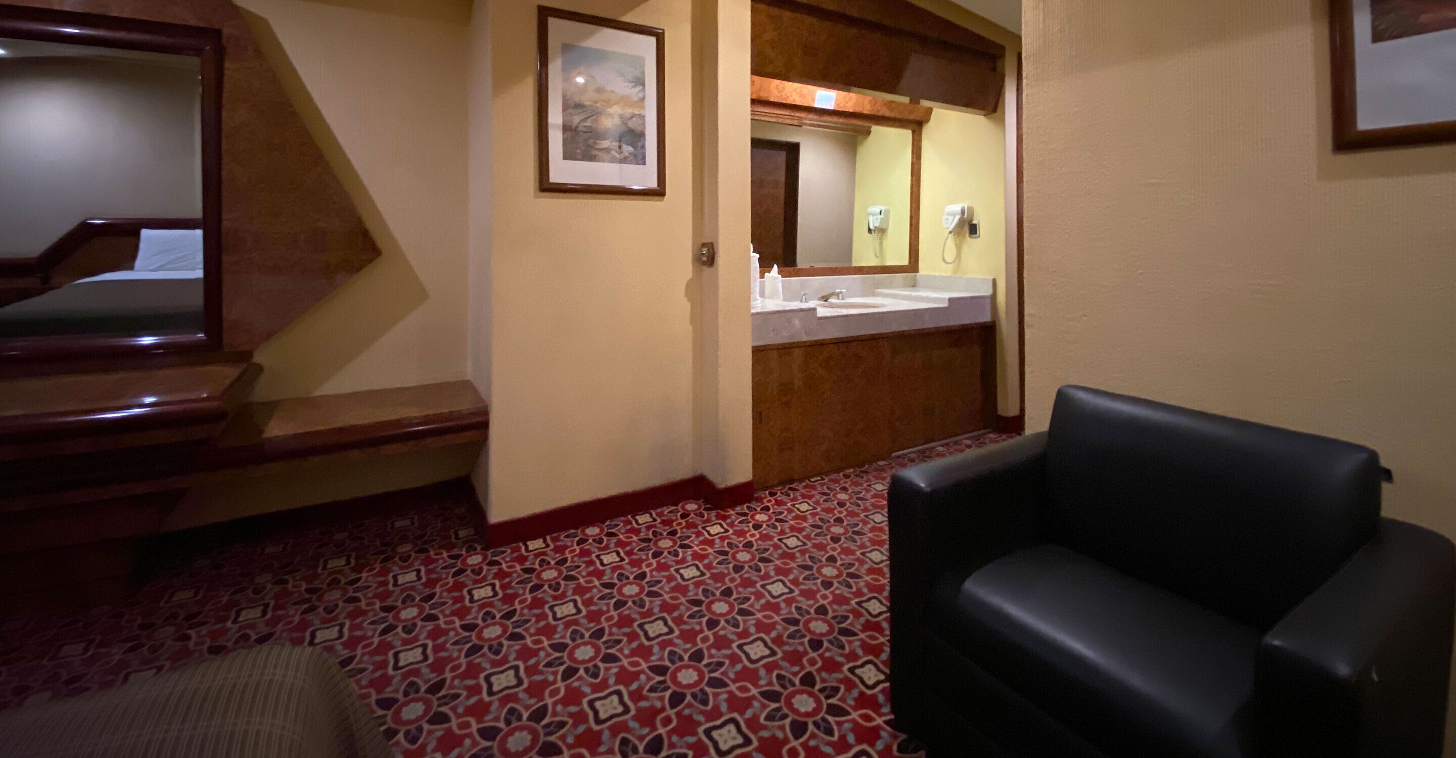 Una habitación de hotel con una escalera de madera, un sillón de cuero negro, una alfombra estampada, un baño con lavamanos y espejo, y un cuadro enmarcado en la pared.