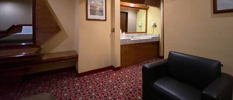 Una habitación de hotel con una escalera de madera, un sillón de cuero negro, una alfombra estampada, un baño con lavamanos y espejo, y un cuadro enmarcado en la pared.