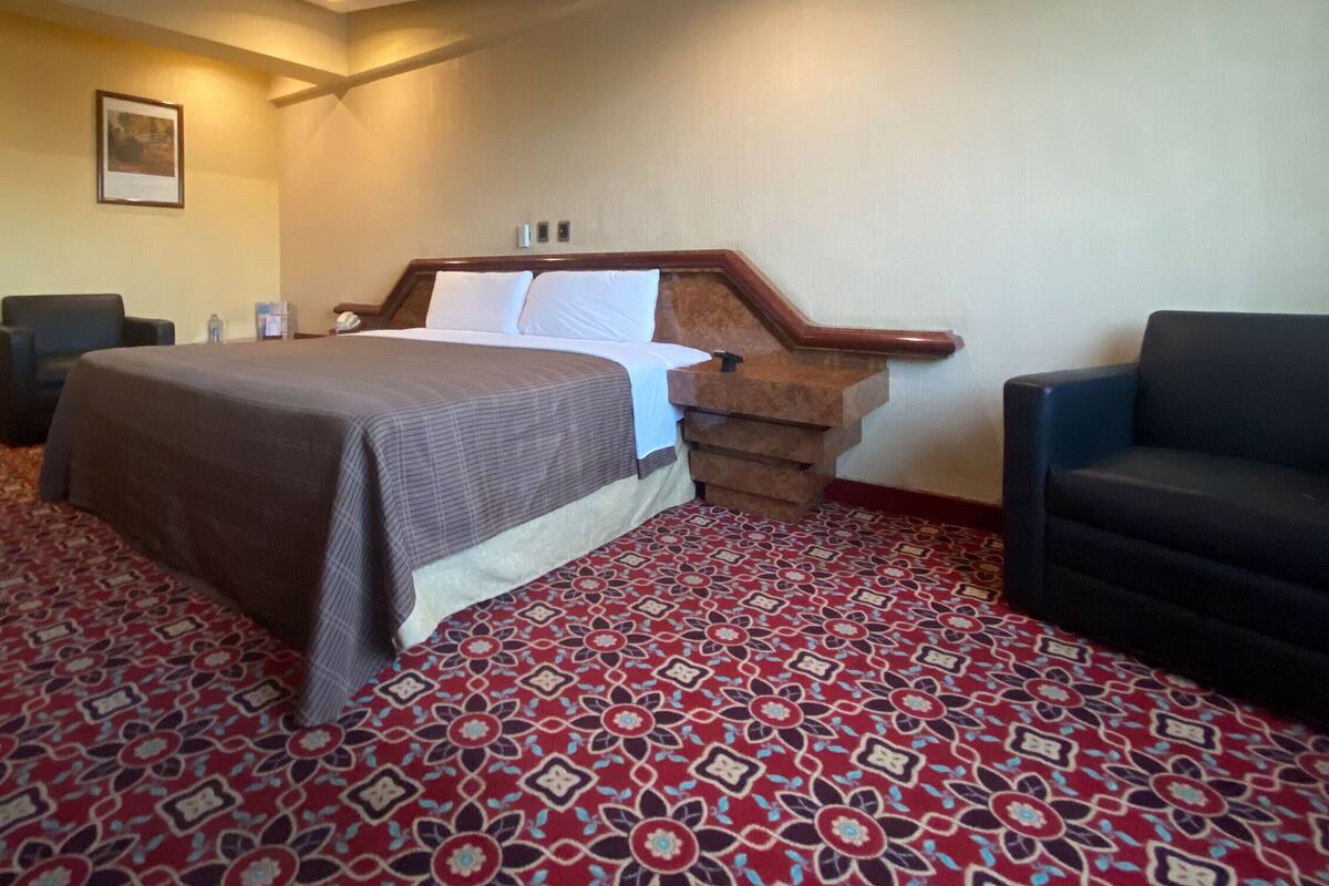 Habitación de hotel con una cama grande, cabecero de madera, alfombra estampada y un rincón de descanso con una silla.