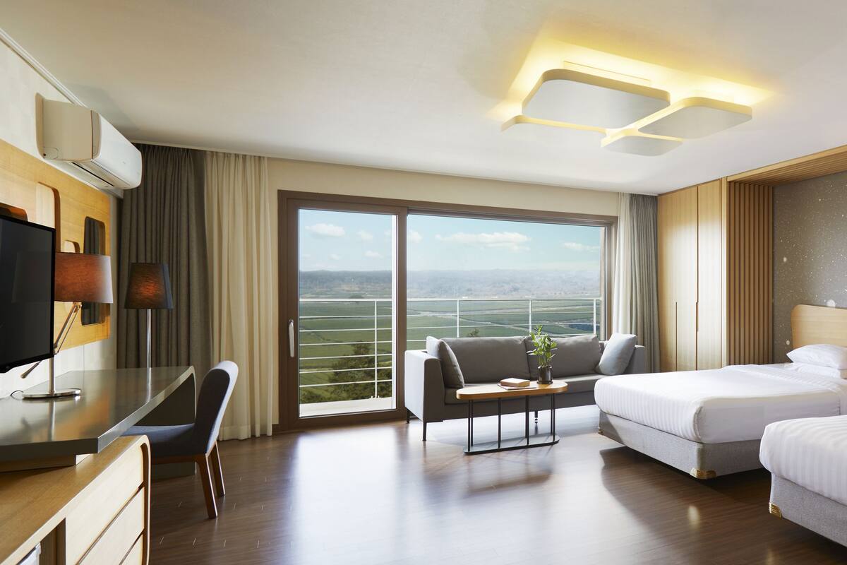 Quarto de hotel com duas camas, uma escrivaninha, uma cadeira, televisão e uma janela grande com vista.