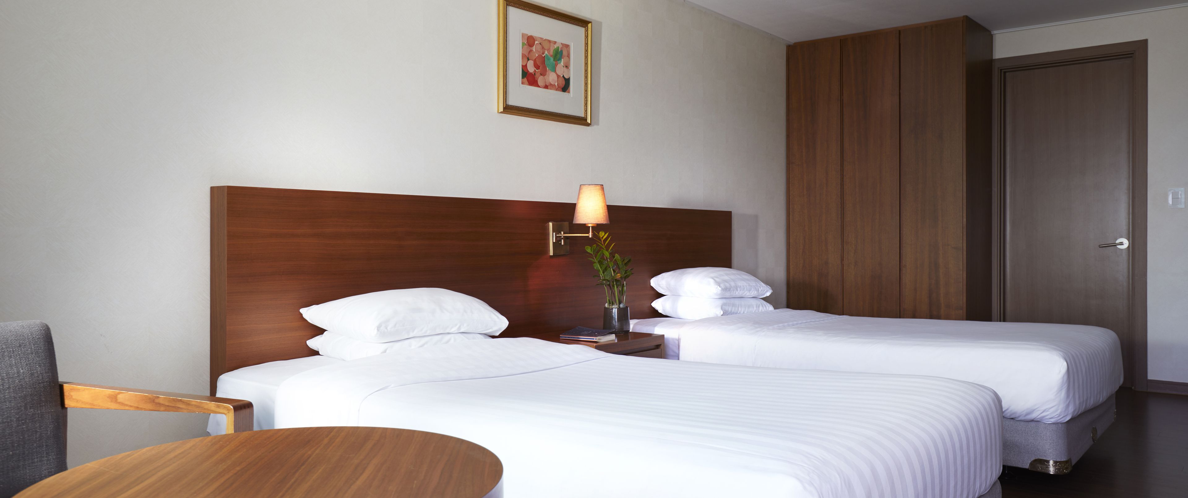 A hotel room with two beds, a wooden headboard, a bedside table with a lamp, a chair, and a small table.