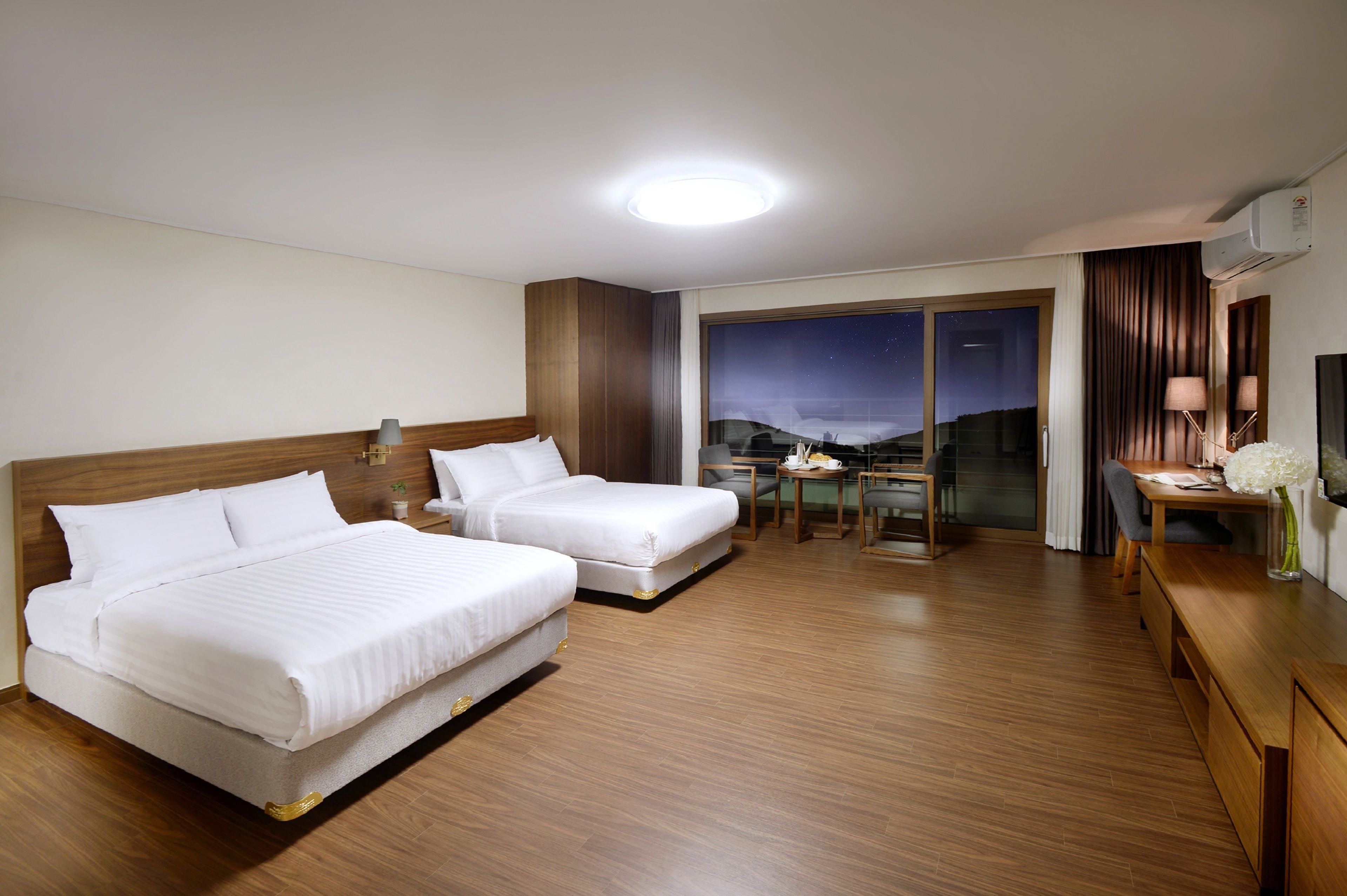 Habitación de hotel con dos camas, un escritorio, una silla, una mesita y vistas al cielo nocturno a través de la ventana.