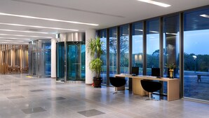 Un espacio de oficina moderno con un mostrador de recepción, dos sillas negras, una planta grande en maceta y vistas a través de amplios ventanales.