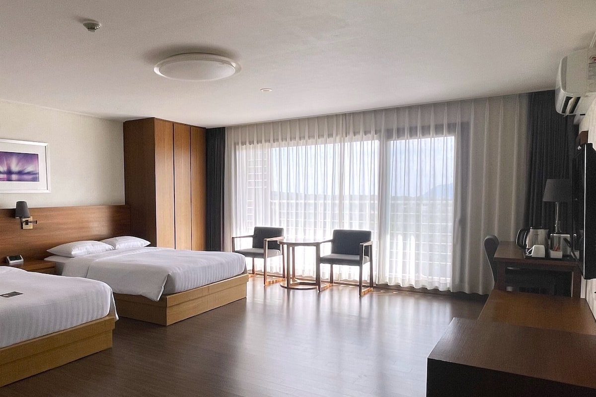 Quarto de hotel com duas camas, uma escrivaninha, cadeiras, cafeteira e uma janela grande com cortinas.