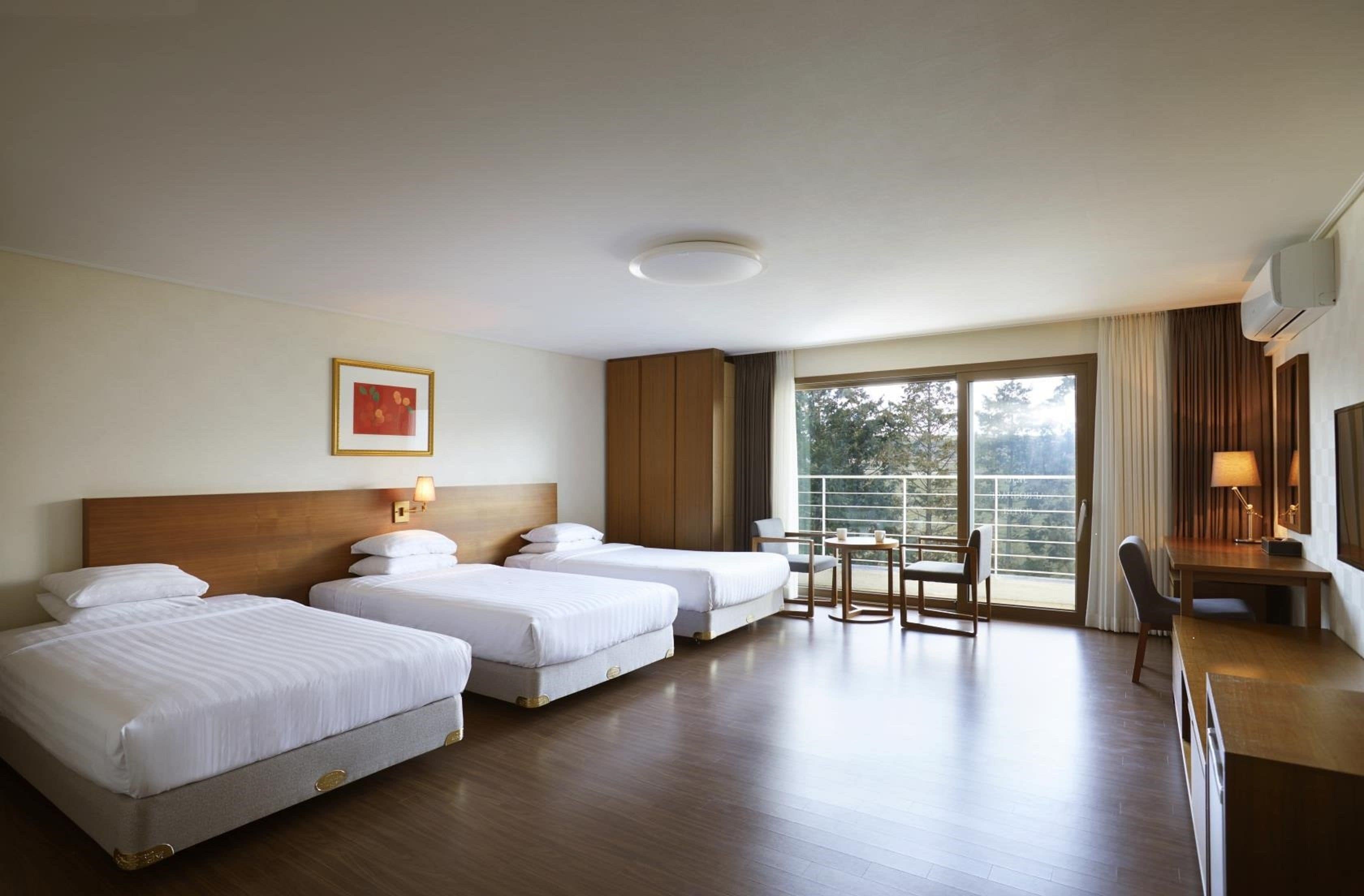 A hotel room with three beds, a desk, a chair, and a large window with a view.