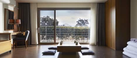 A modern hotel room with a large sliding glass door leading to a balcony, a wooden desk with a chair, a bed with pillows, and a large window.