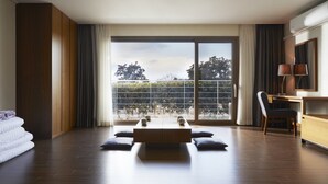 Una habitación de hotel moderna con un gran ventanal corredero a un balcón, un escritorio de madera y una zona de estar acogedora con cojines.