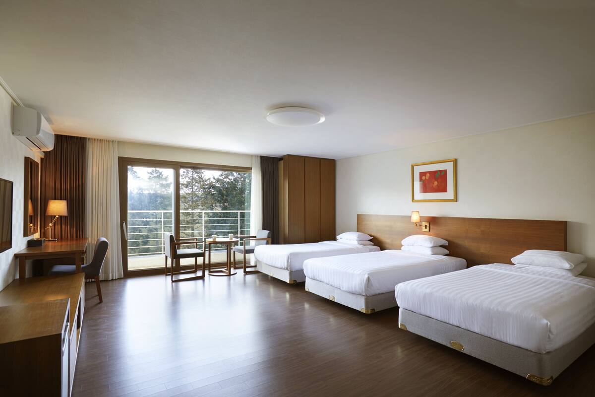 Quarto de hotel com duas camas, uma escrivaninha, uma cadeira, uma mesa e uma janela grande.