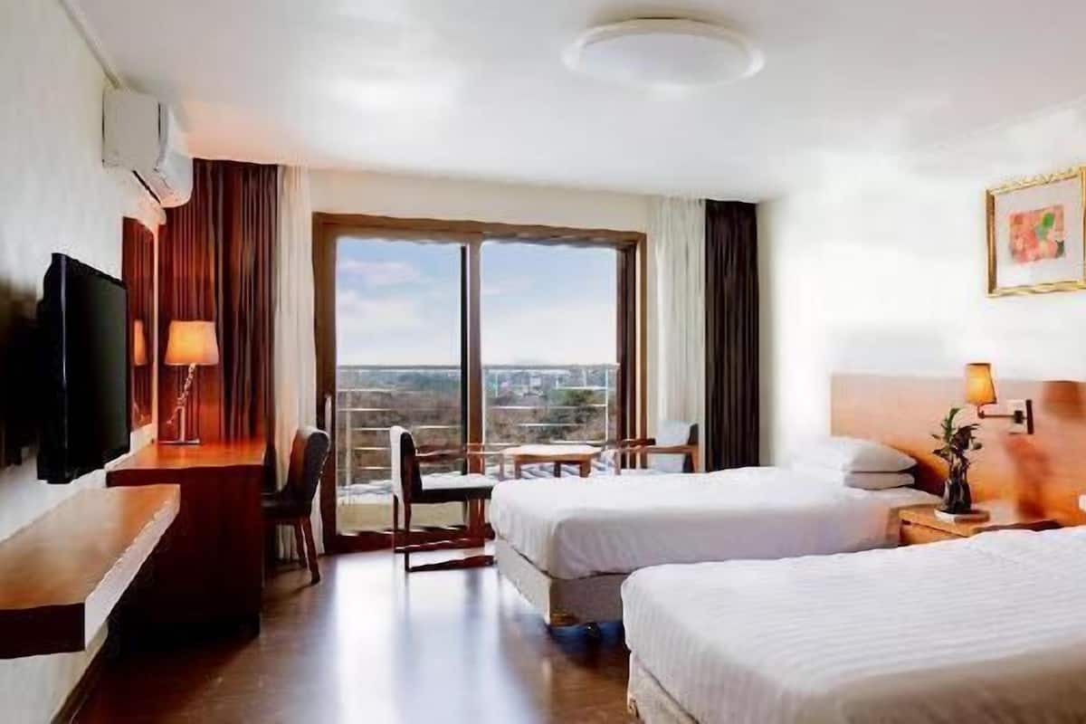 Quarto de hotel com duas camas, televisão, escrivaninha, cadeira, mesa, janela com vista e um quadro na parede.