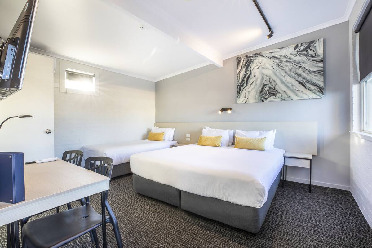 A hotel room with two beds, a desk, chairs, a large marble artwork on the wall, and a window.