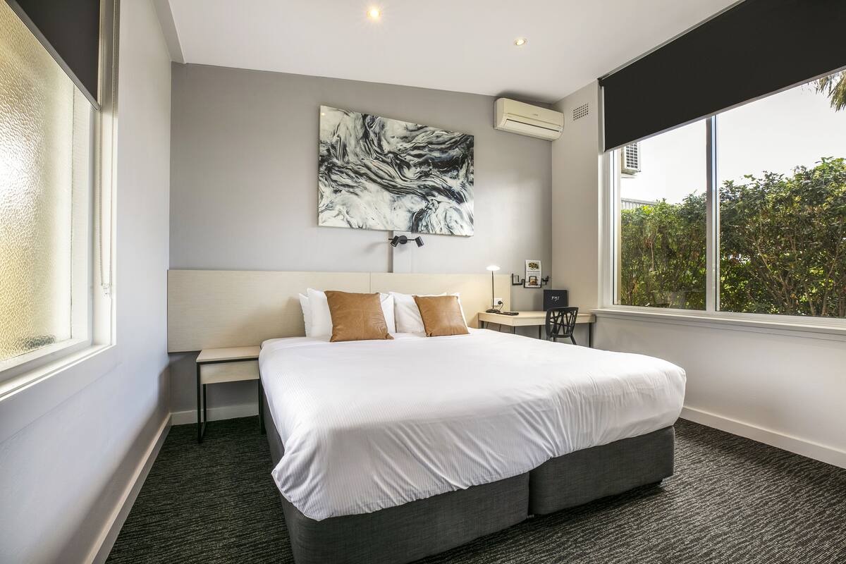 A modern hotel room with a large bed, a desk, and a view of greenery outside.