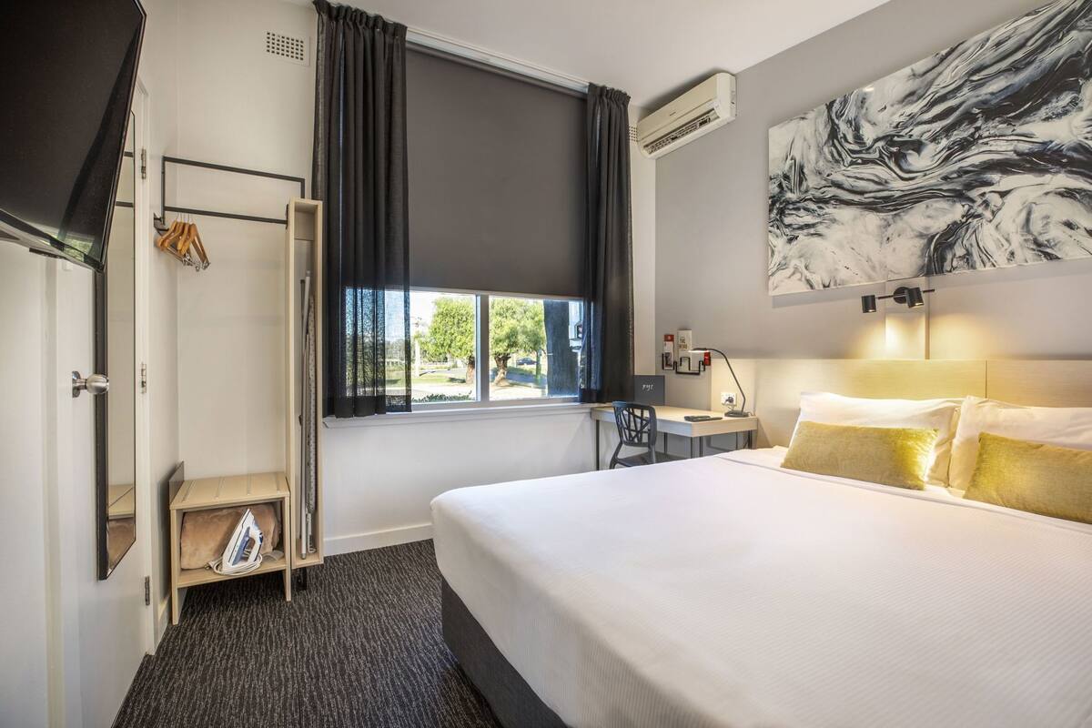 A hotel room with a bed, a desk, a chair, a wardrobe, a window, and an air conditioning unit.