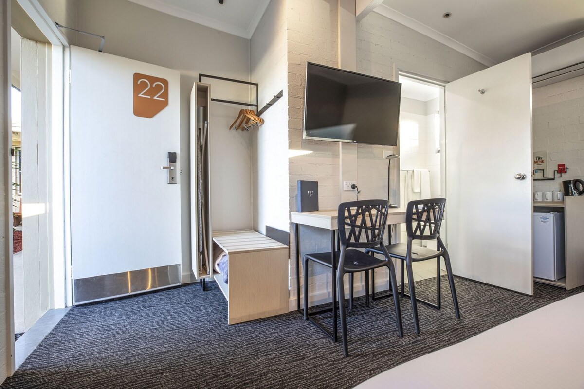 A hotel room with a bed, a desk, two chairs, a TV, a mini-fridge, and a coat rack.