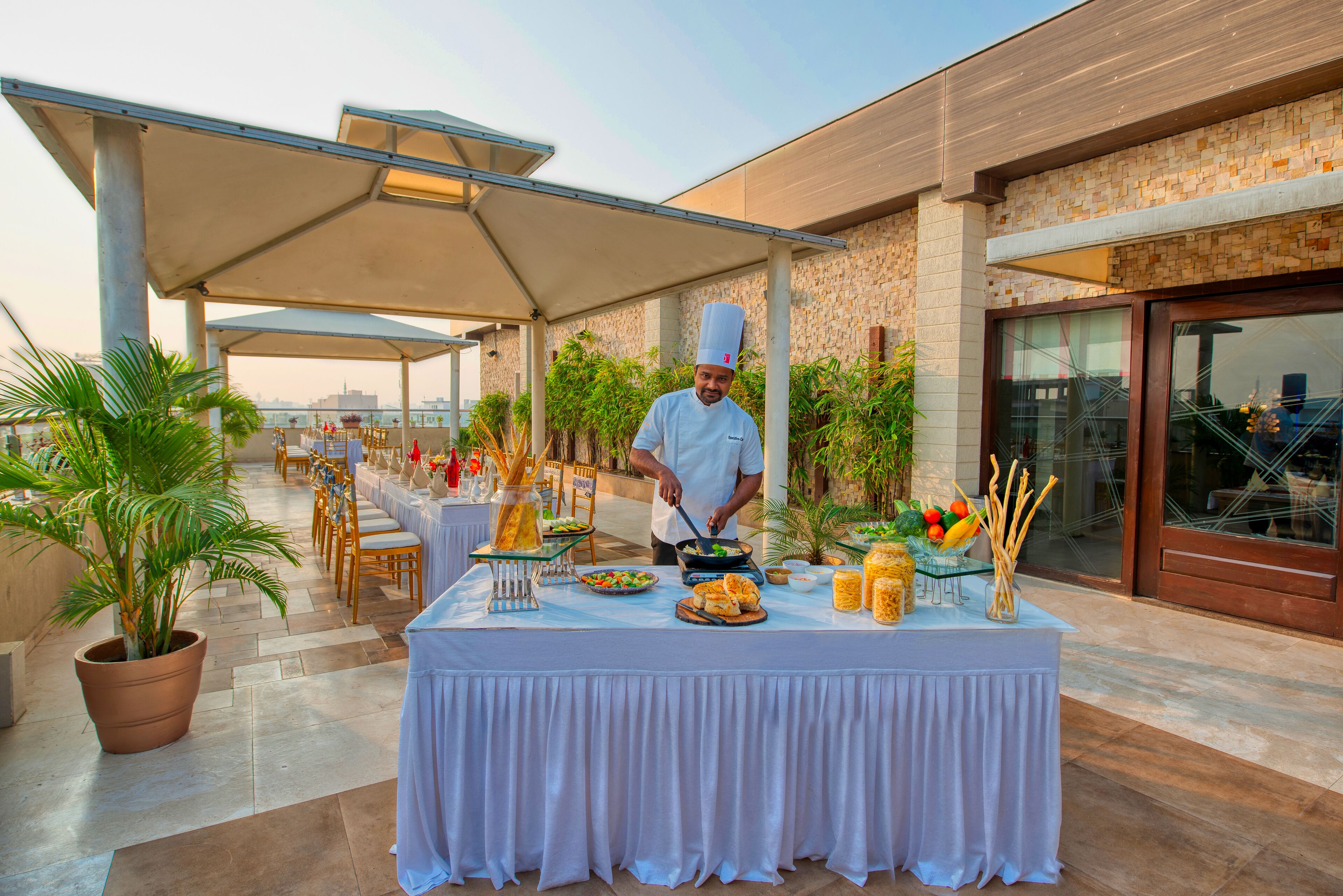 A chef preparing food at an outdoor dining area with a covered table, chairs, and potted plants.