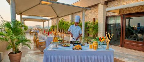 A chef preparing food at an outdoor dining area with a covered table, chairs, and potted plants.