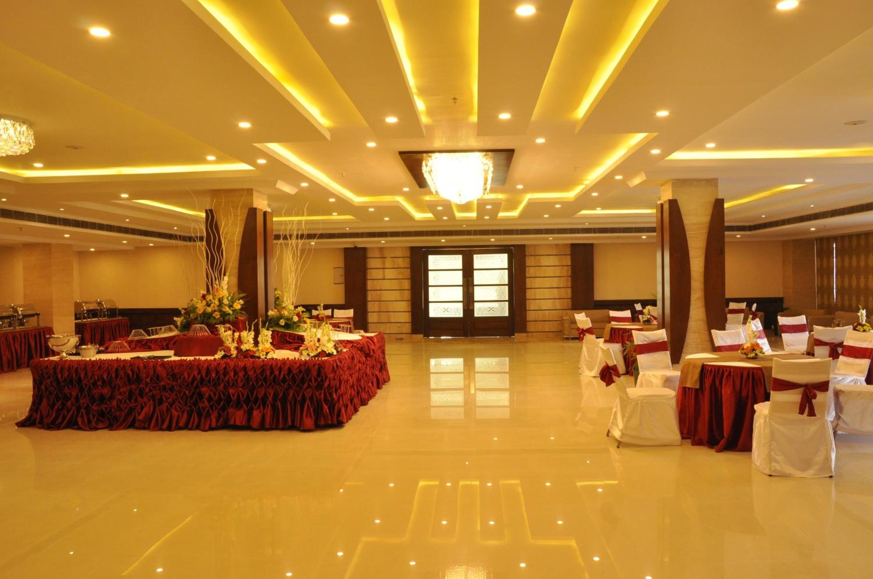 Une salle de banquet spacieuse dotée d’un plancher poli, d’une table ronde centrale agrémentée de compositions florales et de rangées de chaises blanches ornées de rubans rouges.