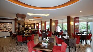 A spacious hotel lobby with a bar area, multiple dining tables, and red upholstered chairs.